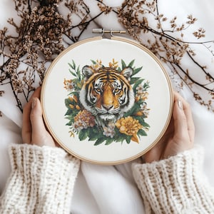Könnte beinhalten: Nahaufnahme eines Tigergesichts, umgeben von gelben und orangefarbenen Blumen. Der Tiger ist in einem Kreuzstich-Stickmuster auf einem weißen Stoff dargestellt.
