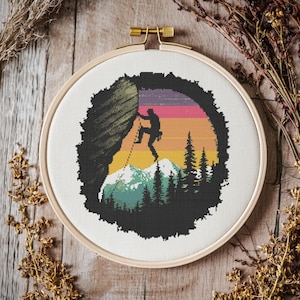 Op de afbeelding: Een kruissteekpatroon met een silhouet van een klimmer die een rotswand beklimt tegen een zonsondergangachtergrond. Het ontwerp bevat een bergketen, bomen en een kleurrijke lucht.