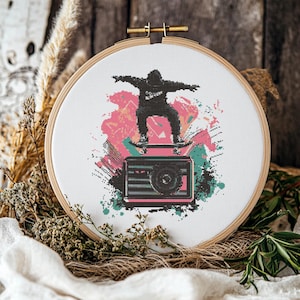 May include: Cross-stitch embroidery hoop art depicting a pixelated skater above a retro camera design. The design uses pink, teal, and black on a white background, framed within a wooden hoop.