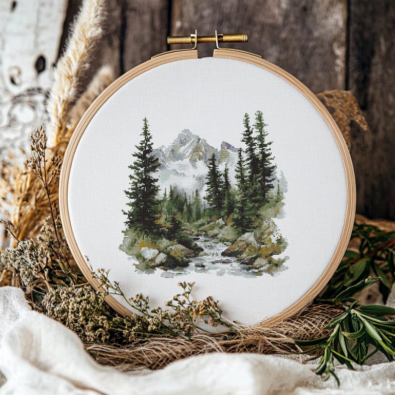 Pode incluir: Um bastidor de bordado em ponto cruz com uma paisagem montanhosa com &aacute;rvores perenes e um riacho. O design utiliza fios verdes, cinzentos e brancos sobre tecido branco. O bastidor de madeira &eacute; vis&iacute;vel. A cena evoca a natureza.