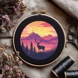 May include: A cross-stitch embroidery hoop featuring a pixelated landscape with a deer silhouette. The design includes mountains, trees, and a sunset in shades of orange, pink, and purple. The hoop is black with a wooden frame.