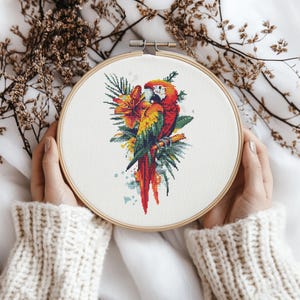 May include: A cross-stitch embroidery hoop with a colorful parrot perched on a branch with tropical flowers and leaves. The parrot is red, yellow, and green with a blue beak.