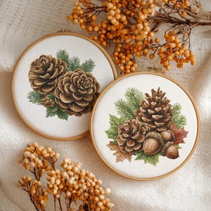 May include: Two round embroidery hoops with detailed cross-stitch designs of pine cones, acorns, and foliage. The designs feature shades of brown, green, and white, set against a neutral background. The hoops are made of light-colored wood.