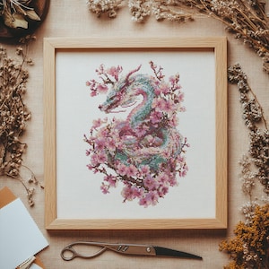 Könnte beinhalten: Kreuzstich-Kunstwerk eines Drachen, umgeben von rosa Kirschblüten, in einem hellen Holzrahmen. Der Drache hat Farbtöne von Türkis, Rosa und Lila. Das Kunstwerk befindet sich vor einem neutralen Hintergrund mit getrockneten Blumen und einer Schere.