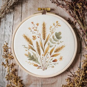 Op de afbeelding: Kruissteekborduurwerk van tarwe en wilde bloemen in een houten hoepel. Het ontwerp heeft tinten goud, groen en bruin, wat een rustieke, natuurlijke uitstraling creëert. De borduurwerk is geplaatst op een crèmekleurige stof.