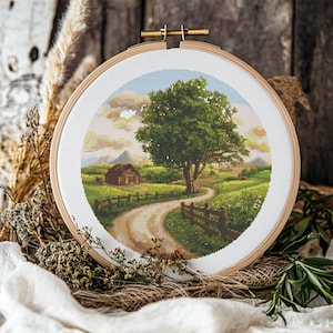 Può includere: Ricamo a punto croce che rappresenta un paesaggio campestre con un sentiero di terra tortuoso che conduce a una piccola casa, un grande albero verde e una recinzione in legno. Il ricamo è incorniciato in un cerchio di legno rotondo.