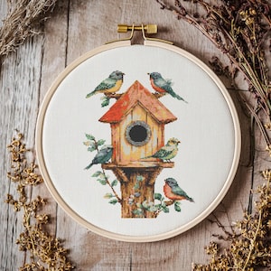 May include: A cross-stitch embroidery of a birdhouse with several colorful birds perched on it. The birdhouse has a red roof and a round opening. The embroidery is set in a wooden hoop.