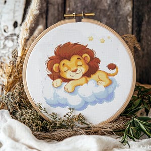 Puede incluir: Un aro de bordado de punto de cruz con un fondo blanco que presenta un león de dibujos animados durmiendo en una nube con estrellas en el fondo.
