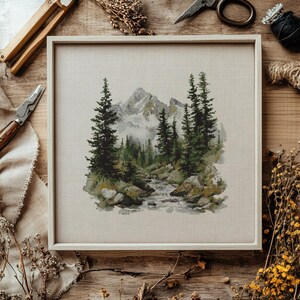 Pode incluir: Uma paisagem em ponto cruz emoldurada com uma cordilheira, &aacute;rvores perenes e um riacho. A obra de arte est&aacute; em uma moldura de madeira clara. A paleta de cores inclui tons de verde, cinza e bege. Ferramentas de artesanato e flores secas circundam a moldura.