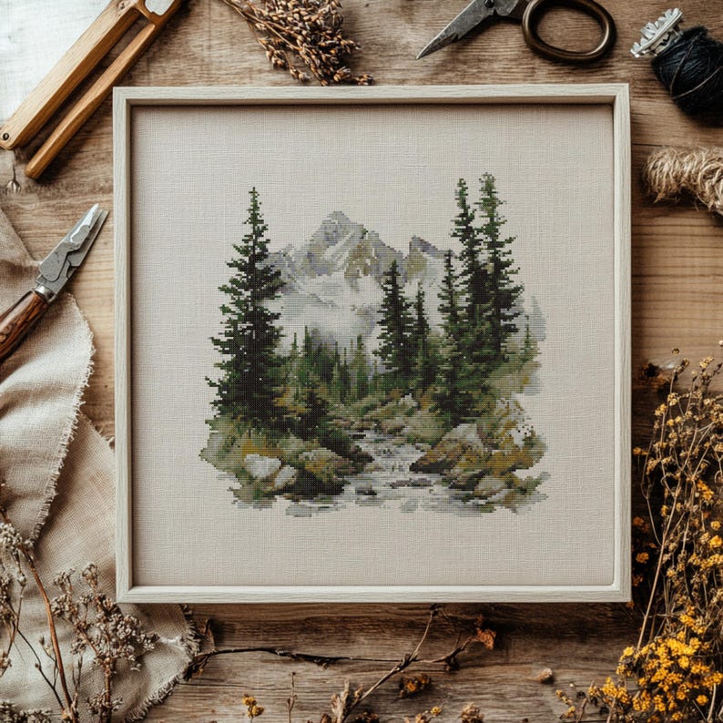 Pode incluir: Uma paisagem em ponto cruz emoldurada com uma cordilheira, &aacute;rvores perenes e um riacho. A obra de arte est&aacute; em uma moldura de madeira clara. A paleta de cores inclui tons de verde, cinza e bege. Ferramentas de artesanato e flores secas circundam a moldura.