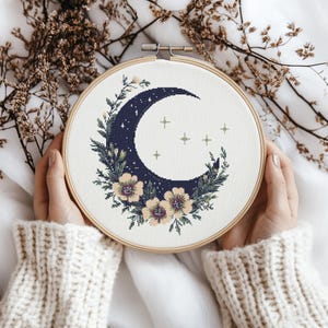 May include: A blue crescent moon with white stars surrounded by a wreath of yellow flowers and green leaves. The moon and flowers are embroidered on a white fabric.