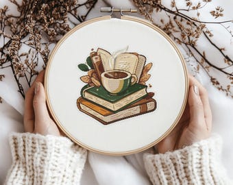 May include: A white embroidery hoop with a cross-stitch design of a cup of coffee and a stack of books. The books are brown, green, and orange. The coffee cup is white with a brown handle and a brown rim. There is a brown leaf on the top of the stack of books.