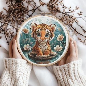 Könnte beinhalten: Ein runder Stickrahmen mit einem farbenfrohen Kreuzstich-Design eines niedlichen Tigerbabys, das auf einem Stein in einem Teich mit Blumen sitzt. Der Tiger hat orange und schwarze Streifen und blaue Augen.