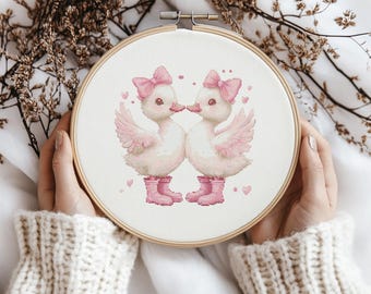 Könnte beinhalten: Ein Kreuzstich-Stickrahmen mit zwei entzückenden Entenküken in rosa Stiefeln und Schleifen, die sich küssen. Das Design beinhaltet rosa Herzen und ist vor einem weißen Hintergrund platziert, gehalten von Händen in einem cremefarbenen Pullover.