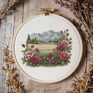 以下が含まれることがあります： 木製のフレームに入ったクロスステッチ刺繍。山、野原、赤いバラのある風景が描かれています。刺繍は白い布の背景に施されています。