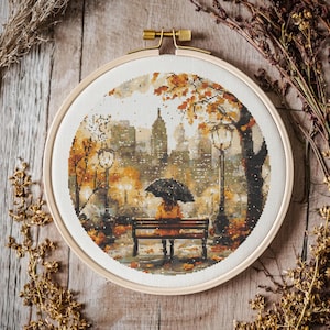 Könnte beinhalten: Eine Kreuzstich-Stickerei in einem runden Holzrahmen zeigt eine Parkszene im Herbst. Das Kunstwerk zeigt eine Person mit einem Regenschirm auf einer Bank, mit Stadtgebäuden im Hintergrund. Die Farbpalette umfasst Orange-, Braun- und Gelbtöne.