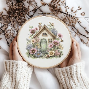 Puede incluir: Un bastidor de bordado con un diseño de punto de cruz de una casa de campo con una puerta verde, rodeada de flores rosas, azules y amarillas. El bordado está sujeto en un aro de madera.