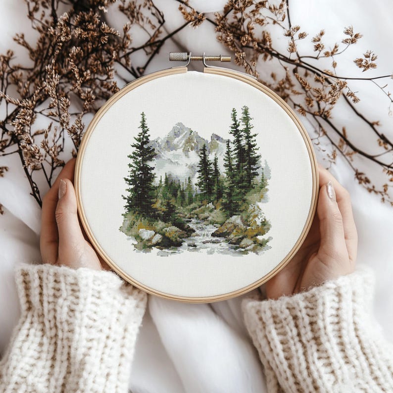 Pode incluir: Um bastidor de bordado redondo com uma cena de paisagem com uma cordilheira, &aacute;rvores perenes e um riacho. A obra de arte &eacute; em tons de verde, cinza e branco, sobre um fundo de tecido branco. O bastidor &eacute; segurado por m&atilde;os que usam um su&eacute;ter creme.