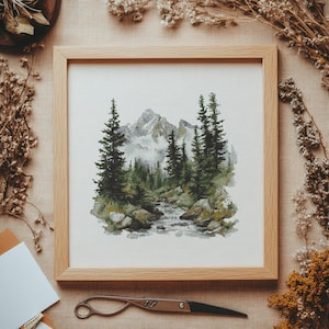 Puede incluir: Obra de arte de punto de cruz enmarcada en madera clara. Representa un paisaje de montaña con árboles verdes, un arroyo y rocas. La paleta de colores incluye verdes, grises y blancos, creando una estética serena y natural.