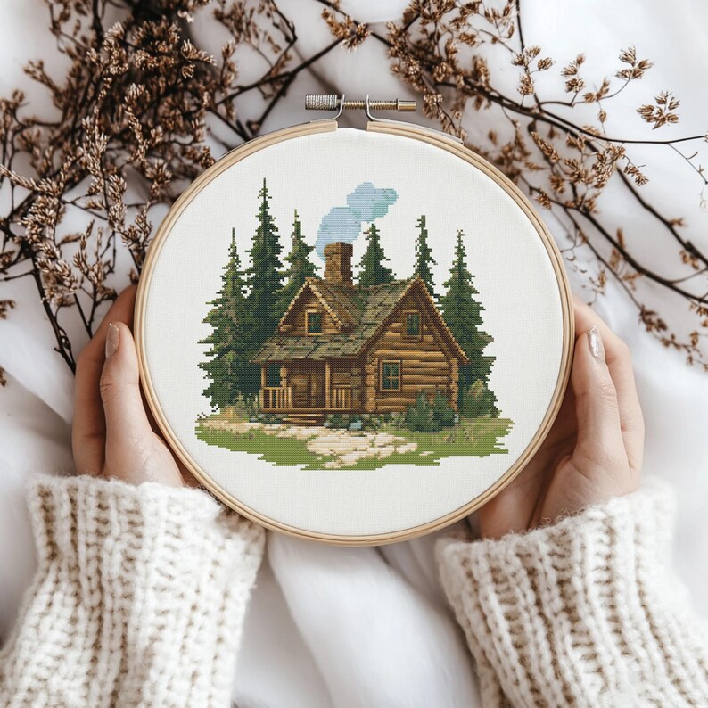 K&ouml;nnte beinhalten: Ein Kreuzstich-Stickrahmen mit einer rustikalen H&uuml;ttenlandschaft. Die H&uuml;tte ist braun mit einem Steinfundament, umgeben von gr&uuml;nen Kiefern und einer blauen Rauchfahne. Die Stickerei wird in einem Holzrahmen gehalten.