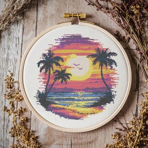 May include: A cross-stitch embroidery hoop with a sunset scene featuring two palm trees, a beach, and a bright orange sun. The colors are vibrant and include shades of pink, orange, yellow, blue, and green.
