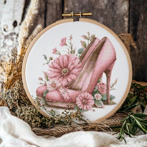 May include: A cross-stitch embroidery hoop depicting a pink high-heeled shoe. The design includes pink flowers and green foliage. The embroidery is displayed in a wooden hoop, suitable for wall decor.
