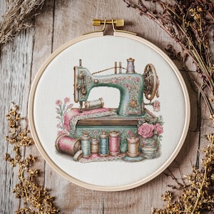 Op de afbeelding: Kruissteekborduurwerk van een vintage naaimachine in een houten hoepel. De machine is turquoise met bloemendetails, omringd door roze en turquoise garenklosjes en roze rozen. De achtergrond is een houten oppervlak.