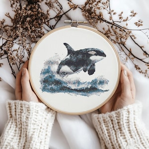 May include: A cross-stitch embroidery hoop showing a black and white orca leaping from blue and white waves. The orca is the central image, set against a white backdrop. The embroidery is framed in a wooden hoop.