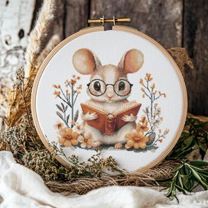 May include: Cross-stitch embroidery of a mouse wearing glasses and reading a book, surrounded by flowers. The design is set in a wooden embroidery hoop. The mouse is beige and white, the book is brown, and the flowers are orange.