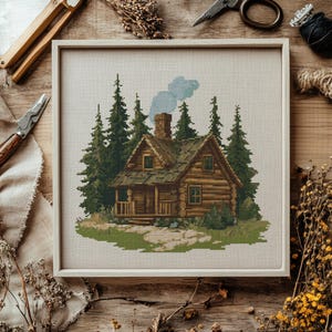 K&ouml;nnte beinhalten: Ein gerahmtes Kreuzstichbild eines rustikalen Blockhauses inmitten gr&uuml;ner Kiefern. Das Haus hat eine braune Holzfassade, einen Schornstein mit blauem Rauch und eine kleine Veranda. Das Kunstwerk ist vor einem neutralen Stoffhintergrund.