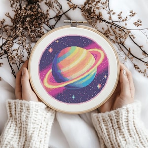 May include: A cross-stitch embroidery of a planet with rainbow stripes and rings, set against a starry purple background. The embroidery is framed in a wooden hoop. The craft project is held by a person wearing a cream-coloured sweater.
