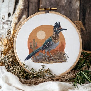 May include: A cross-stitch embroidery hoop featuring a roadrunner bird in a desert landscape. The bird has blue, brown, and black feathers, set against a backdrop of a setting sun, cacti, and desert plants. The hoop is made of wood.