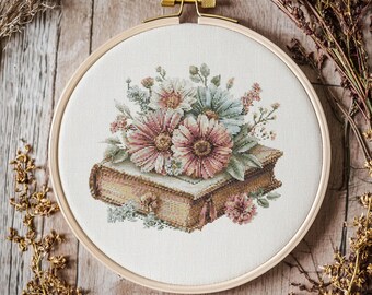 May include: A cross-stitch embroidery of a book with flowers in a wooden hoop. The book is tan with pink and white flowers on top. The hoop is made of light wood with a gold-colored tightening mechanism.