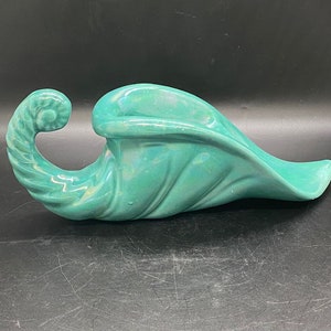 May include: A teal ceramic cornucopia vase with a curled handle. The vase is glazed with a mottled finish.