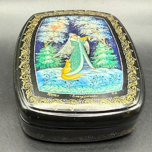 May include: Black lacquered box with a hand-painted winter scene. The lid features a woman in a teal dress and a golden fox. The scene is set against a blue sky with trees and snow. The box has a gold filigree border.