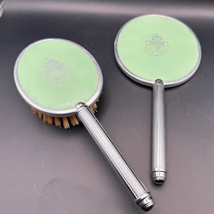 May include: A set of two vintage hand mirrors with silver handles and green mirrored surfaces. Each mirror has a decorative design in the center.