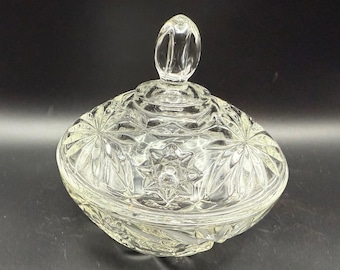 May include: Clear glass candy dish with lid. The dish features a starburst and leaf pattern. The lid has a loop handle. The dish is round and sits on a dark surface.