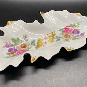 May include: A white porcelain leaf-shaped serving dish with a gold rim and a floral design. The flowers are pink, white, yellow, and purple.