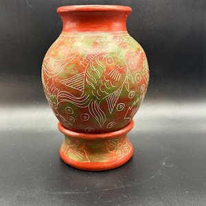 May include: A handcrafted, red and green ceramic vase with fish and floral designs. The vase has a rounded body and a flared rim, sitting on a matching pedestal base. The fish are outlined in white, adding contrast to the earthy tones.