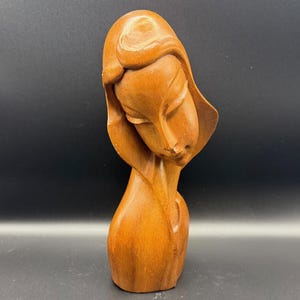 May include: A wooden sculpture of a woman's head and shoulders, carved from light brown wood. The woman's head is tilted downward, with her eyes closed. Her hair is styled in a simple, elegant fashion. The sculpture has a smooth, polished finish.