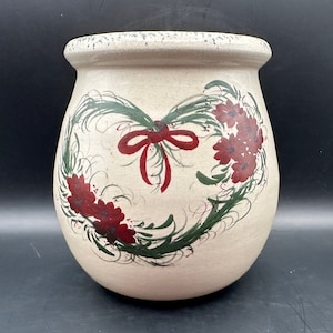 May include: A cream-colored ceramic pot with a heart-shaped design featuring a red bow and green leaves with red flowers.
