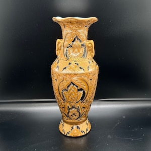 May include: A gold and black ceramic vase with intricate floral designs. The vase has two handles and a wide mouth.