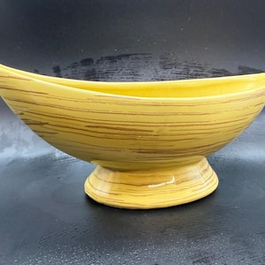 May include: A yellow ceramic bowl with a boat-like shape, featuring brown horizontal stripes. The bowl sits on a rounded base. The interior is a solid yellow color. This decorative piece is suitable for serving or display.