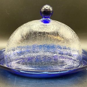 May include: A cobalt blue glass butter dish with a domed, crackle-textured lid and a matching circular base. The lid is topped with a small, round, dark blue knob. The dish is designed to hold and display butter.