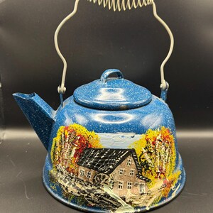 May include: A blue enamel tea kettle with a white speckled finish, featuring a hand-painted scene of a house with a water wheel surrounded by yellow and orange autumn trees. It has a metal handle with a coiled wire grip and a matching lid.