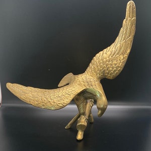 May include: A gold-colored brass eagle sculpture with its wings spread wide. The eagle is perched on a base with its talons extended.