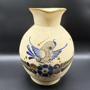 May include: A cream-colored ceramic pitcher with a speckled texture, featuring a hand-painted blue bird and a dark blue floral design with green leaves around the base. The vessel has a wide, rounded body and a flared, open spout at the top.