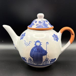 May include: White ceramic teapot with blue floral and folk art designs. The teapot features a blue figure with a staff, a blue lid with floral patterns, and an orange handle. A charming addition to any kitchen.