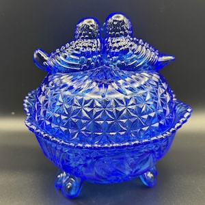 May include: A cobalt blue glass trinket dish with a lid, featuring two birds perched atop. The dish has a geometric pattern and sits on three small feet. The overall design is ornate and vintage.