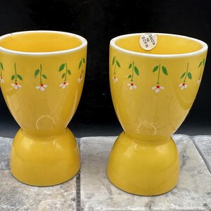 May include: Two yellow ceramic egg cups with white and green floral designs. The cups are made in Korea.